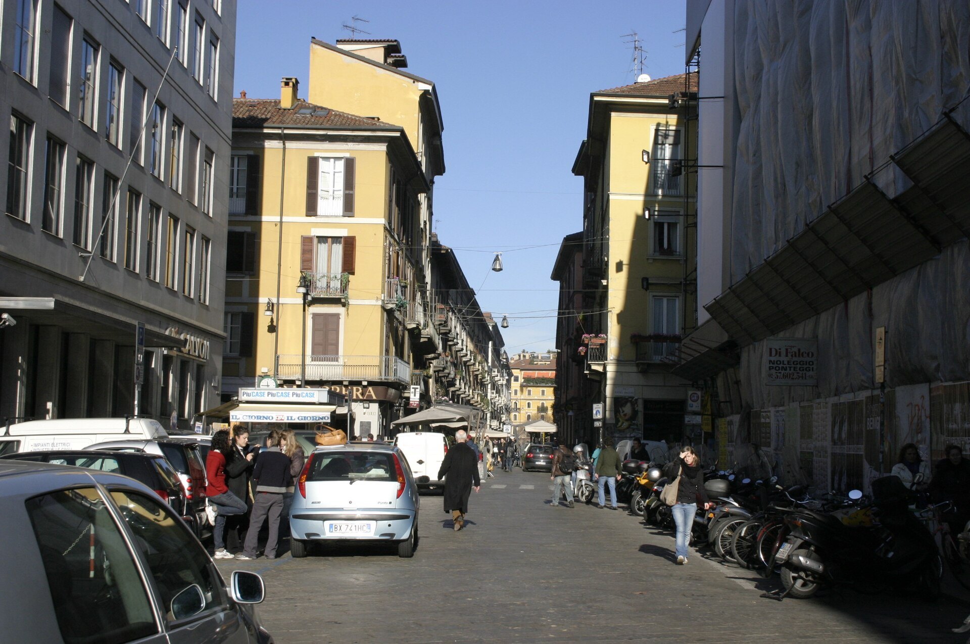 Via Brera with parked cars and scooters and people walking in the middle of the road