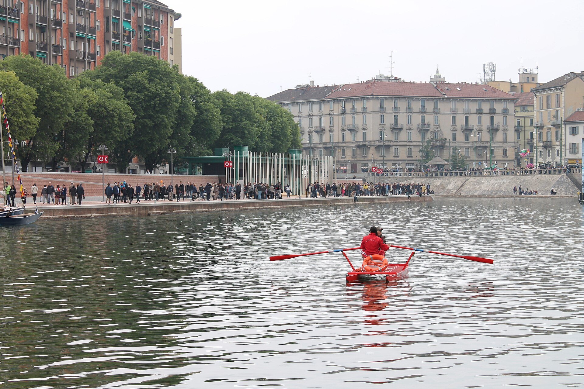 A single sculler rowing on the Darsena with people walking the quays behind