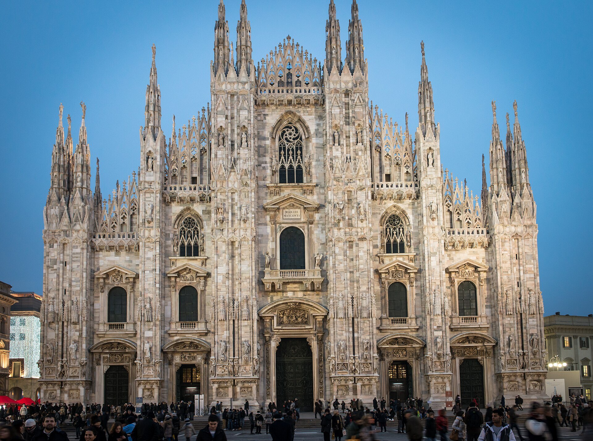 The full marble front of Milan Cathedral under a blue sky, its spires and statues in rows