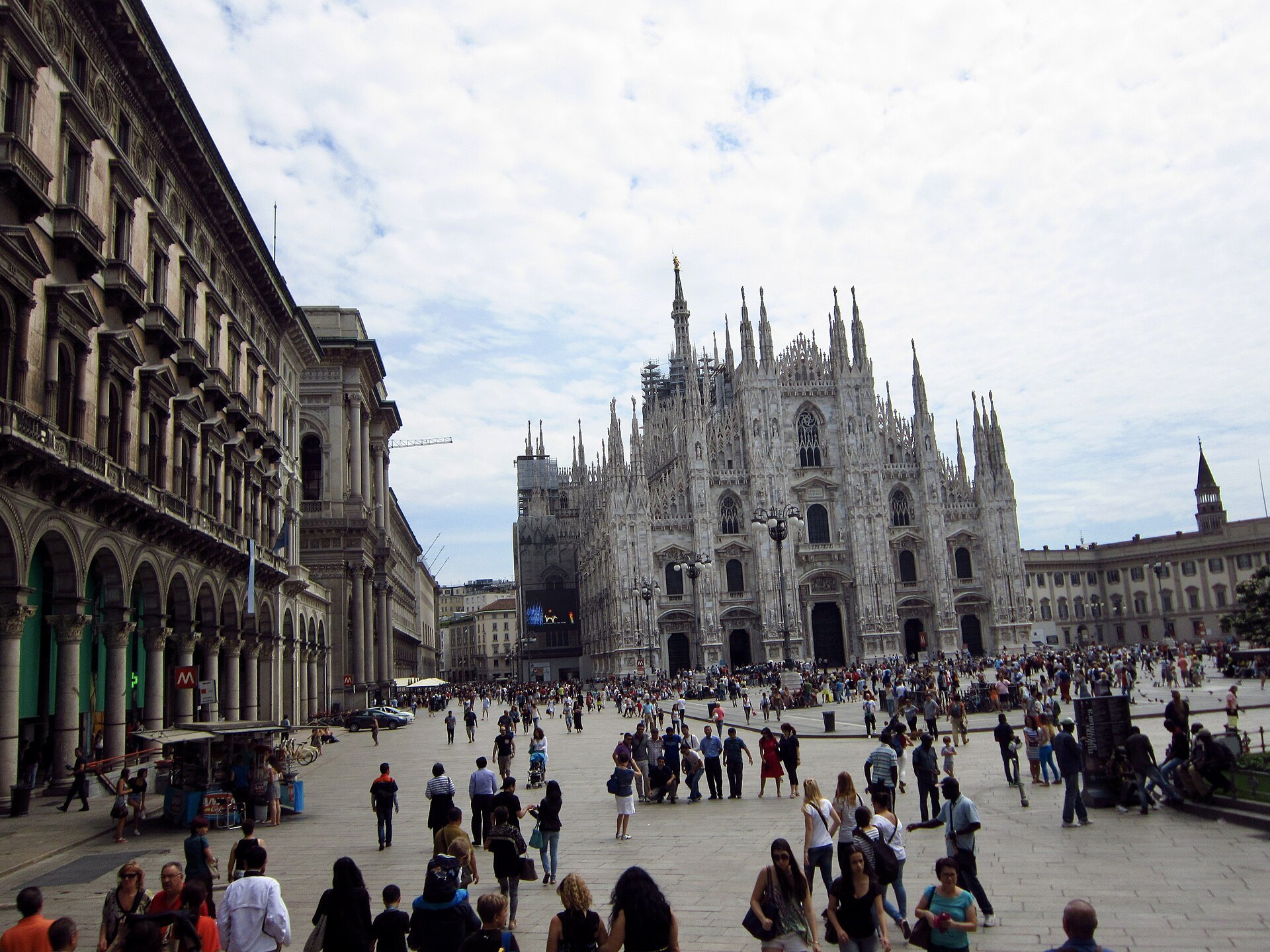 The square seen along the arcade, crowds crossing towards the cathedral at the end of it