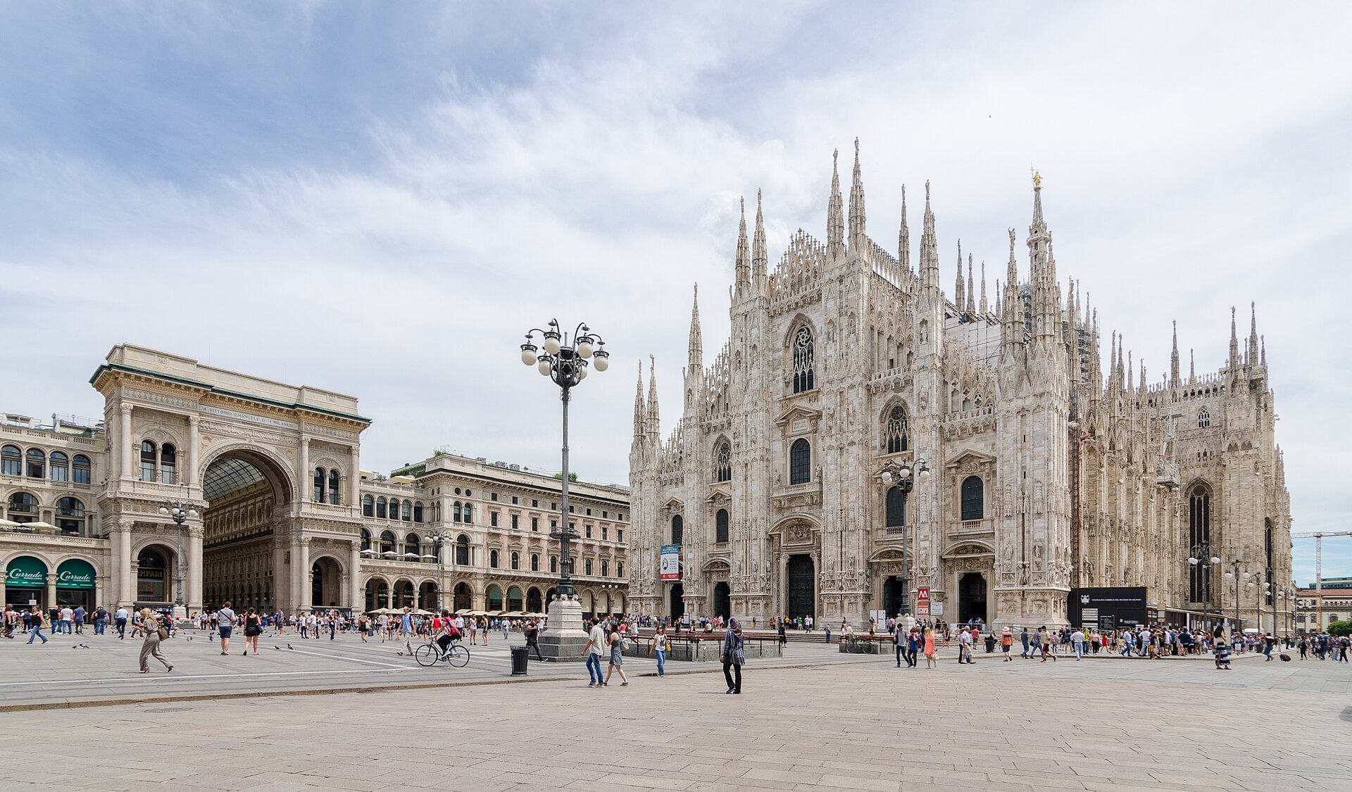 Piazza del Duomo in Milan, the cathedral's marble front on the right and the arched entrance to the Galleria on the left, with people crossing the square between them